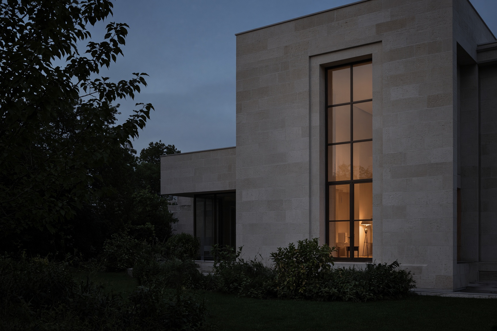 A stone and glass residence at blue hour, one warm window in a dark garden.